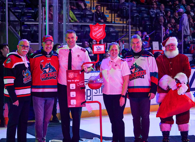 Group of people standing on an indoor sports rink beside a red Salvation Army donation kettle on a stand, with a donation sign and hockey rink boards and seating visible in the background.