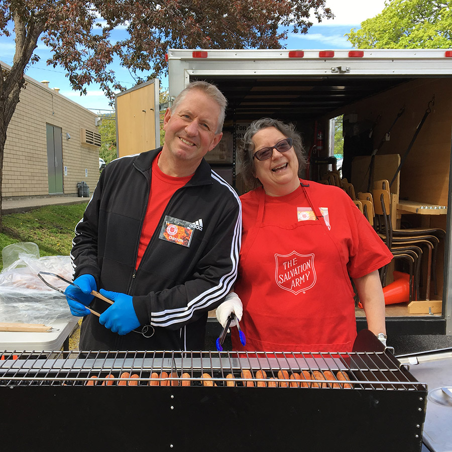 Two volunteers wearing Salvation Army shirts stand beside an open trailer filled with stacked chairs, preparing or organizing items outdoors next to a building.
