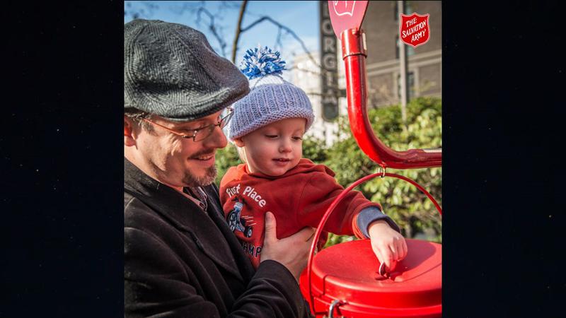 Adult holding a young child next to a red Salvation Army donation kettle, as the child places a coin into the slot, with the kettle stand and Salvation Army shield visible outdoors.