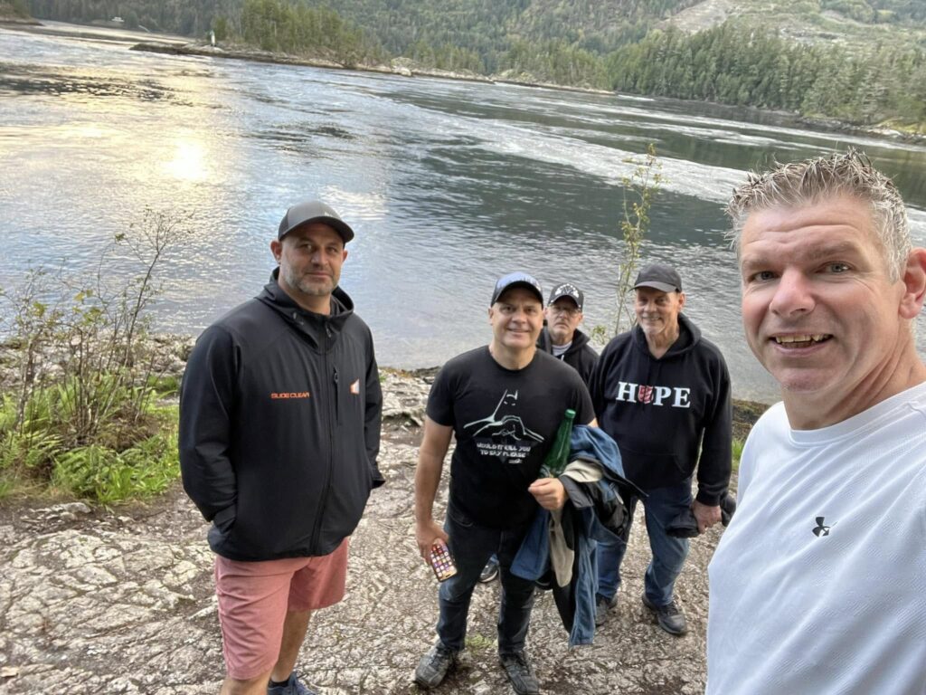 Group of men standing together on a rocky shoreline beside a calm body of water, with trees and greenery nearby, posing for a photo during an outdoor men’s ministry activity.