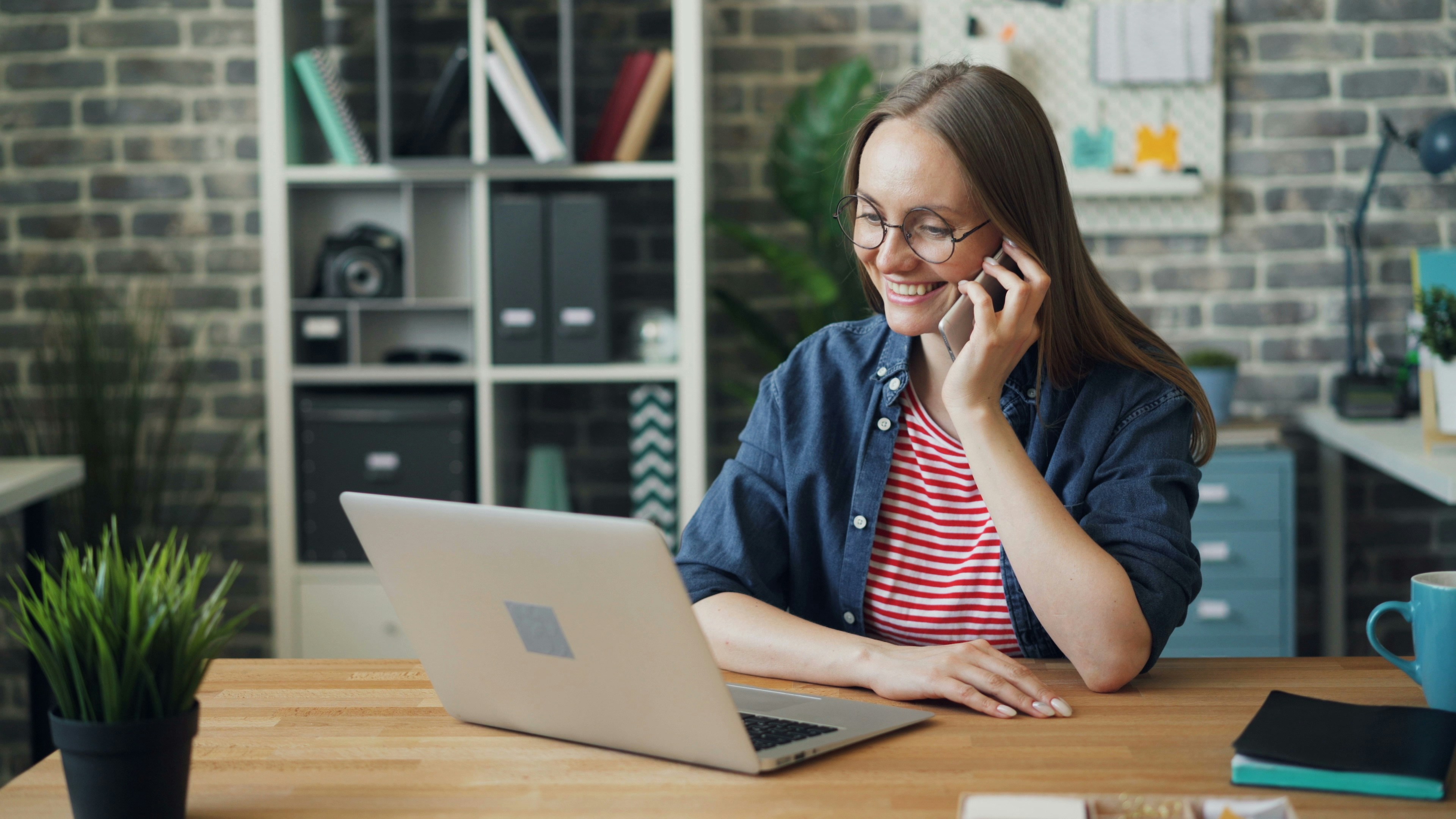 Person seated at a desk using a laptop while holding a smartphone to their ear.