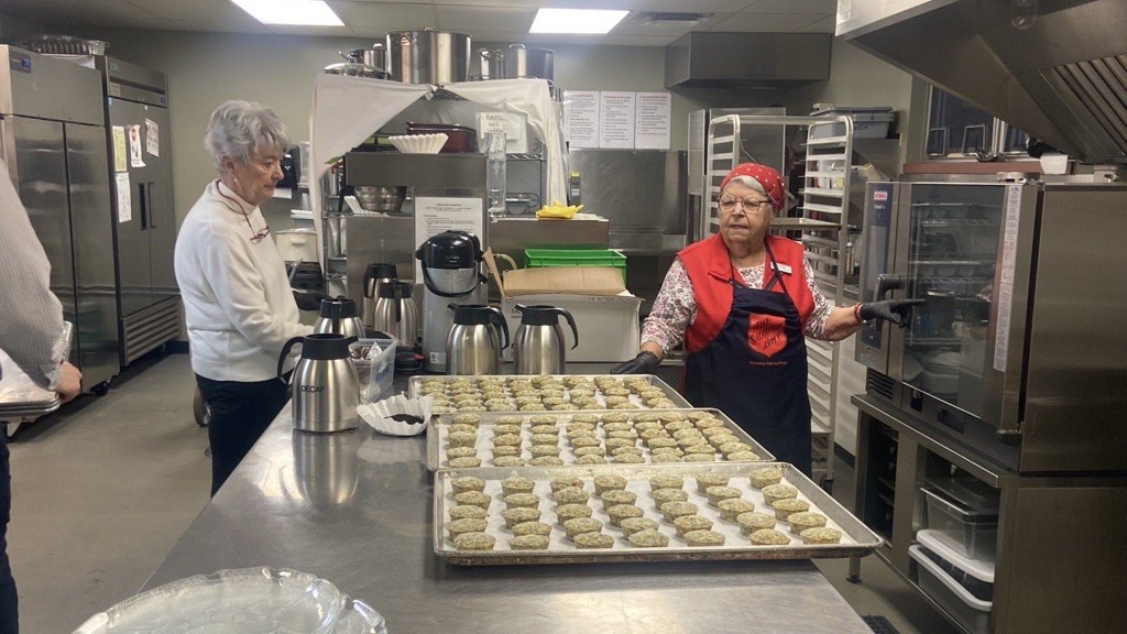 Seniors working together in a commercial kitchen, preparing trays of baked goods on a stainless steel counter alongside coffee carafes and kitchen equipment.