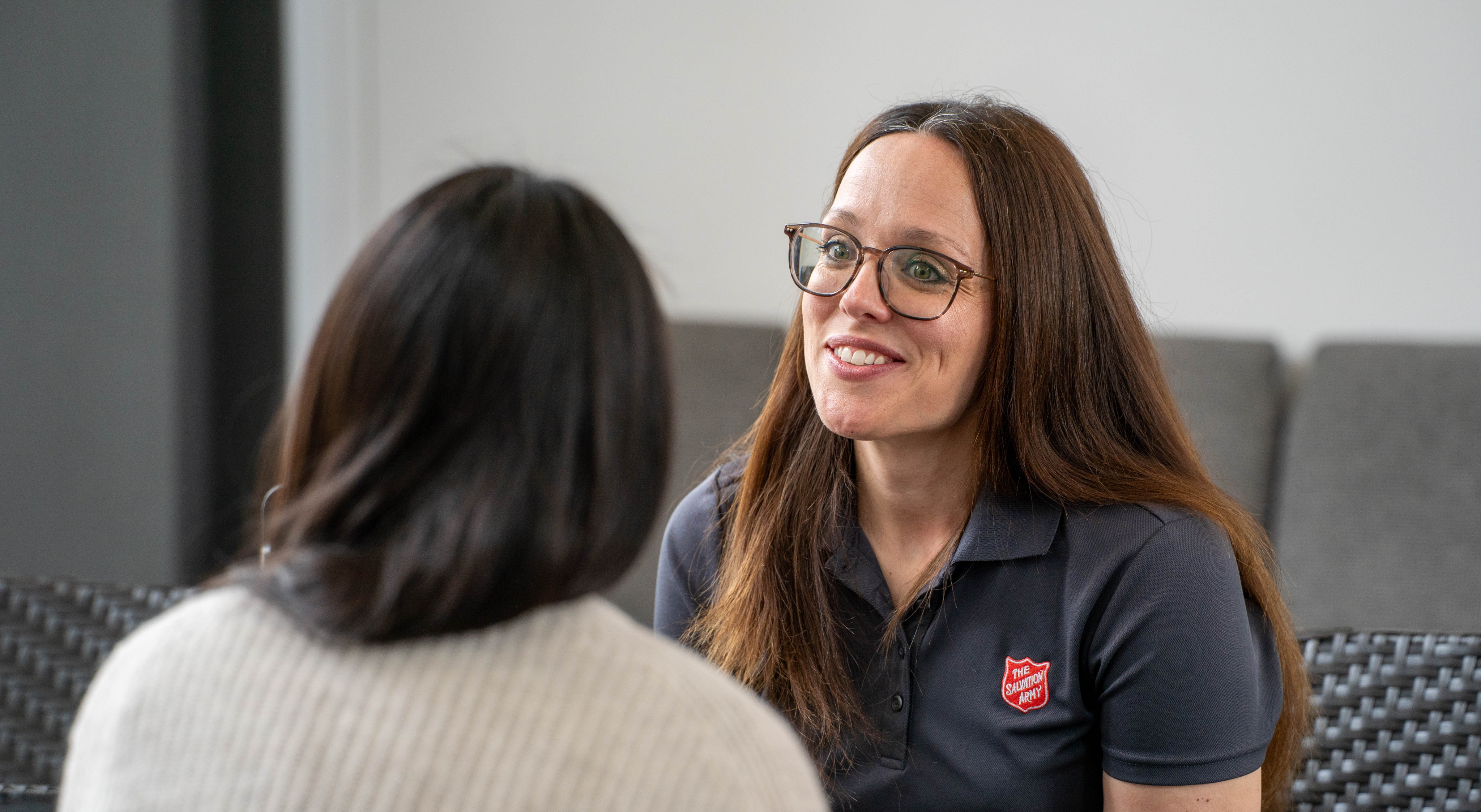 Two people seated indoors facing each other during a one-on-one meeting, with one person wearing a Salvation Army logo shirt, representing a caseworker providing community assistance in a private conversation setting.