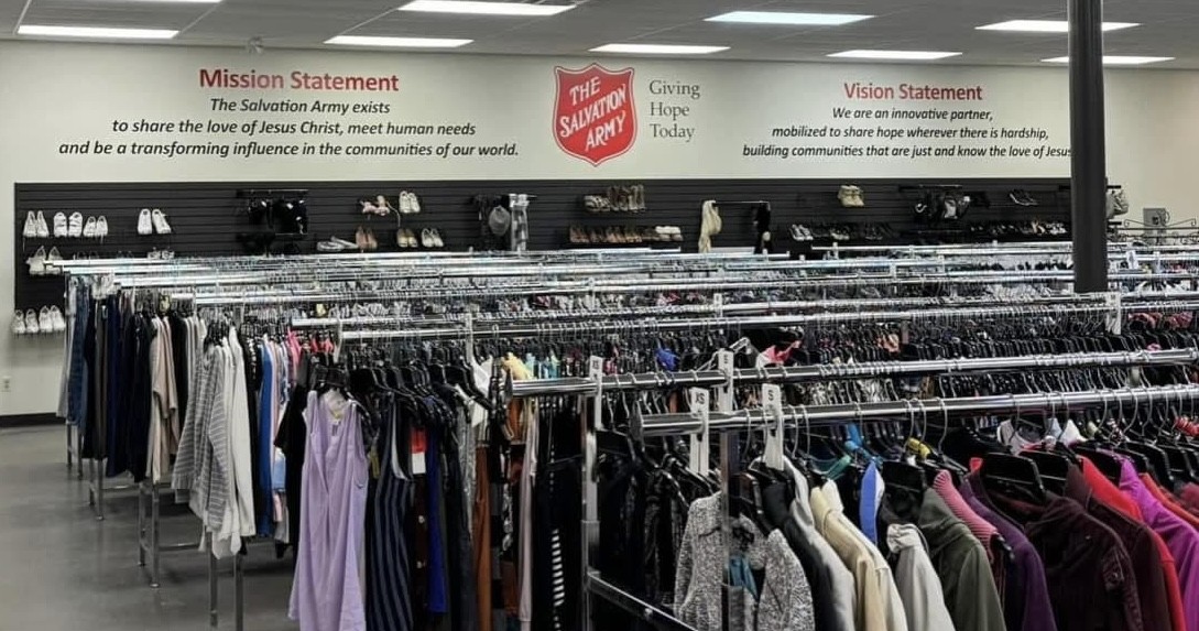 Interior of a Salvation Army thrift store with multiple rows of clothing racks filled with assorted garments, and shelves displaying shoes along the wall beneath signage with the organizations mission and vision statements.