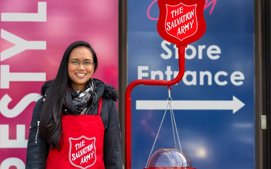 Kettle volunteer standing beside a red Salvation Army donation kettle and stand near a store entrance, wearing a Salvation Army apron.