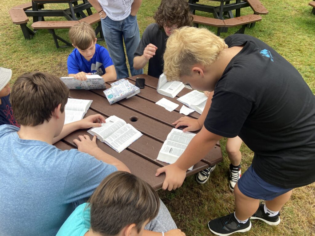 Children and youth gathered around an outdoor picnic table reading and discussing from open books, with additional books and materials spread across the table during a group activity.